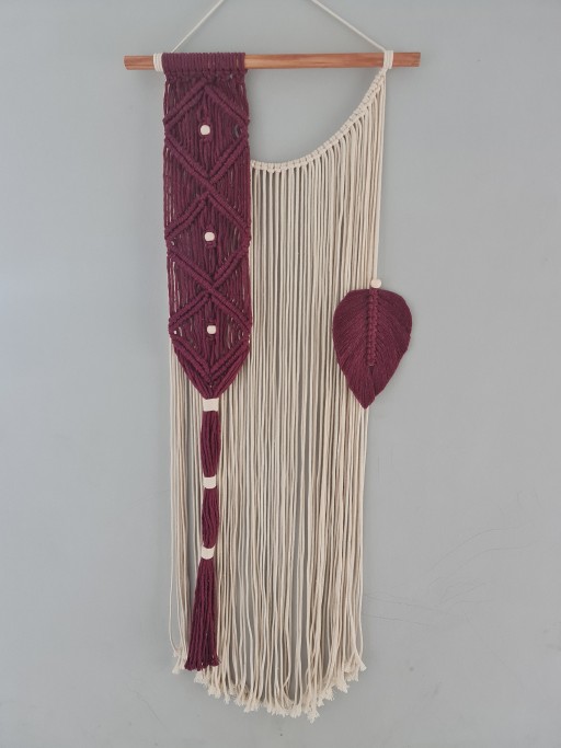 Apresenta um elegante padrão em macramê na cor vinho, complementado por detalhes em contas de madeira e uma charmosa folha decorativa na mesma tonalidade, destacando-se sobre a base de cordões em tom cru.
