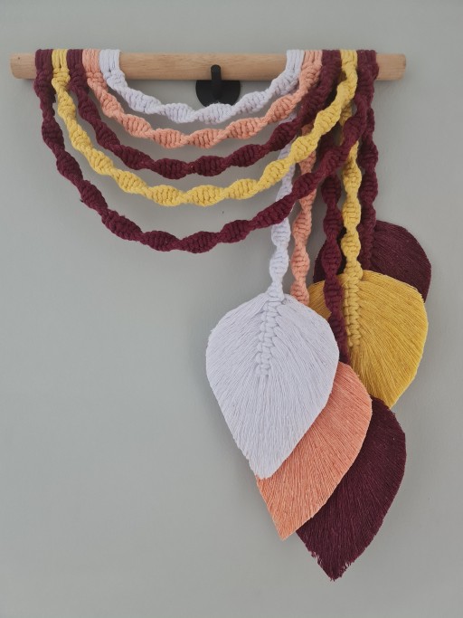 Guirlanda torcida em macramê com folhas pendentes, combinando tons terrosos como branco, pêssego, mostarda e bordeaux. Feita com fios duráveis e suporte de madeira resistente, é perfeita para enriquecer qualquer decoração.